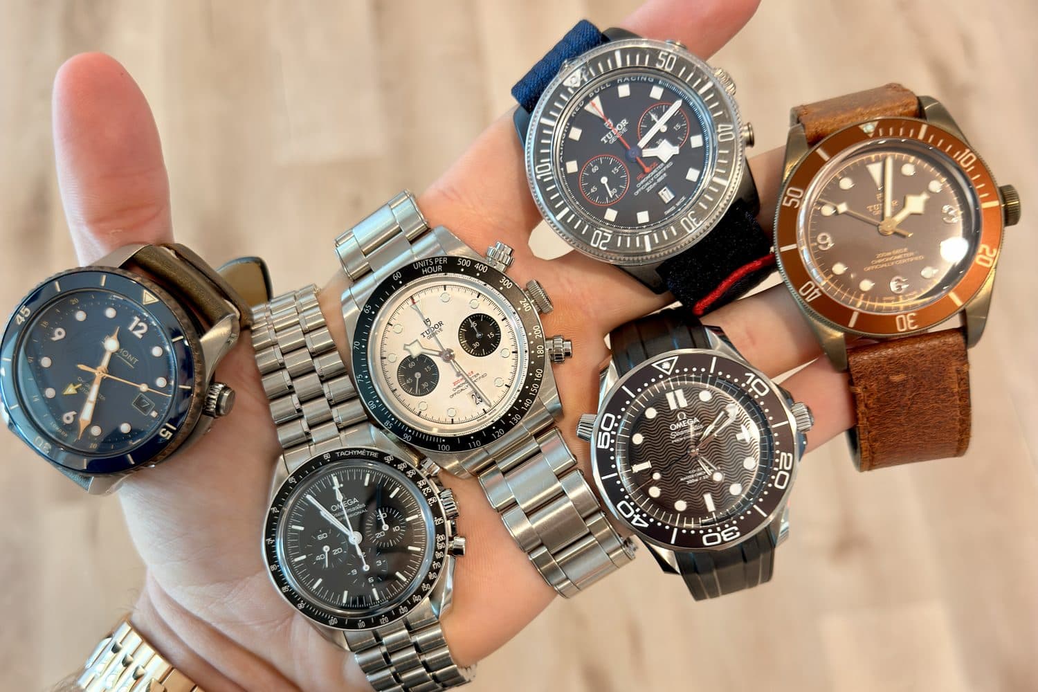 The six starting watches stacked on one hand, seen from above: a Bremont, an Omega Speedmaster, a Tudor Black Bay Chrono, a Tudor Pelagos, an Omega Seamaster and a bronze Tudor Black Bay