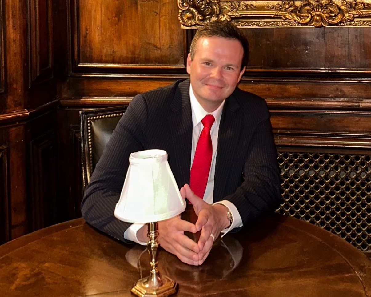 Vadim, founder of Moda, in a dark suit and red tie, seated at a round wooden table in a wood panelled room