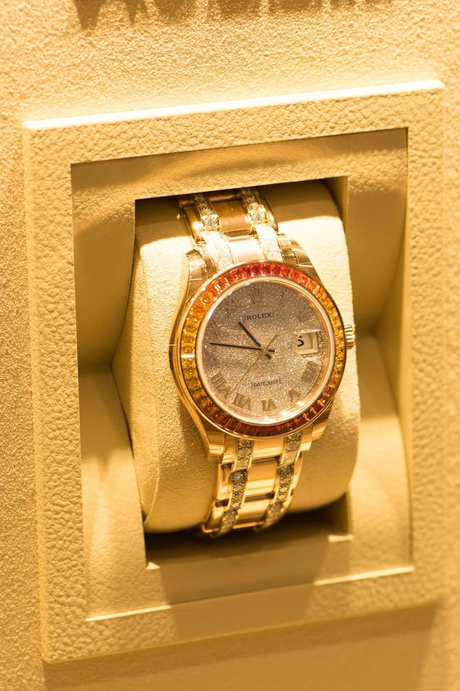 A gold Rolex with a rainbow-coloured sapphire bezel and diamond-paved dial on a Pearlmaster bracelet with diamond-set links, in a display case
