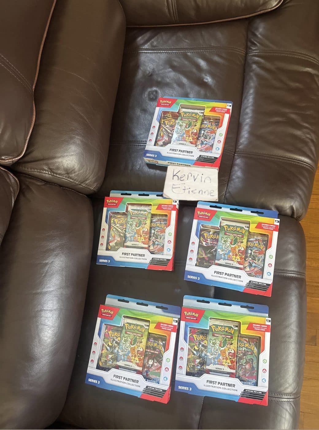 Five sealed Pokemon First Partner Illustration Collection boxes laid out on a leather couch with Kervin Etienne's handwritten name card