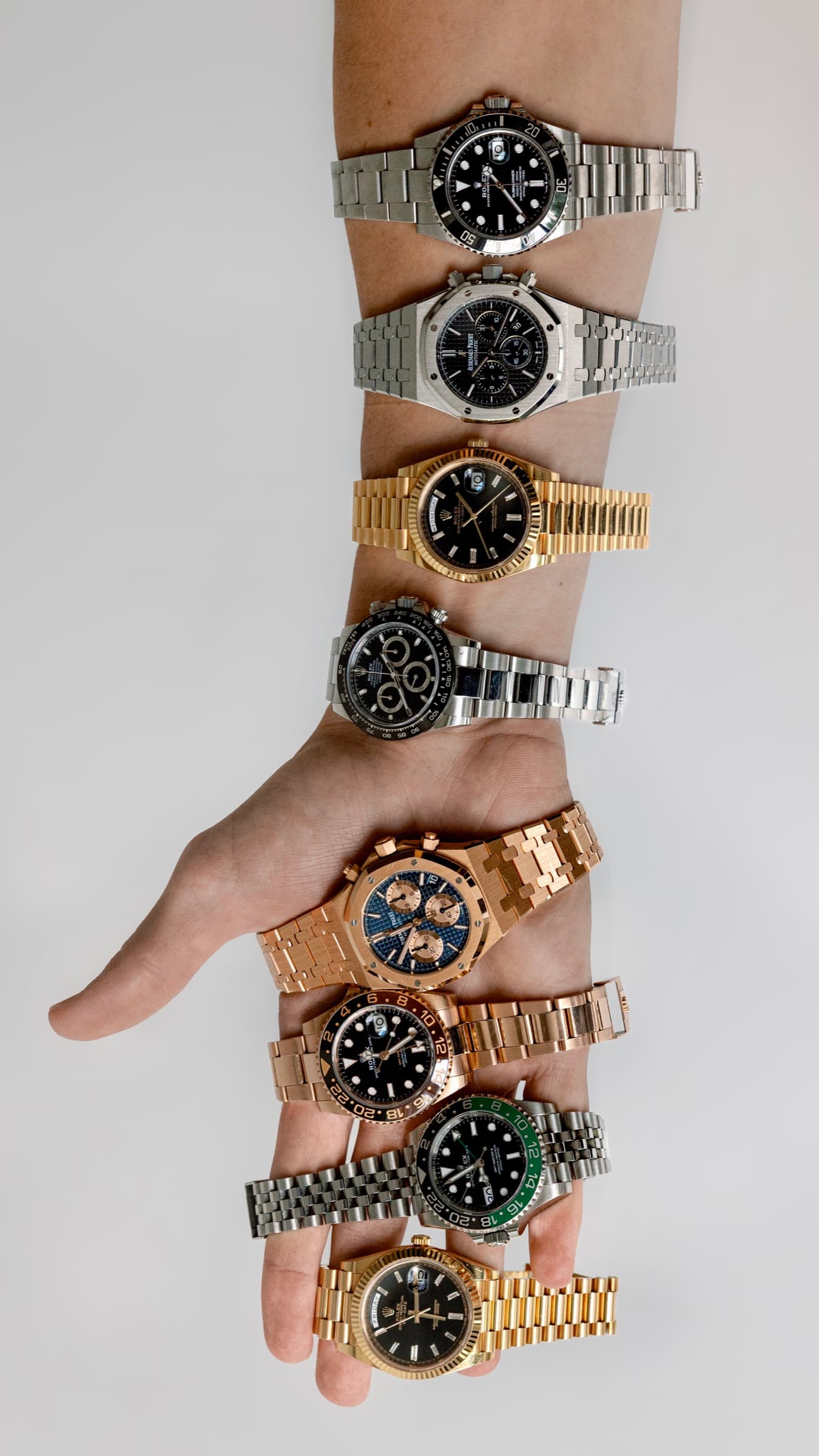 A wrist showcasing several luxury watches lined up in a row, including a Rolex Daytona, Audemars Piguet Royal Oak, and a yellow gold Day-Date. Real Watch Buyers (RWB) is the premium private watch trading service of Moda Clubs.